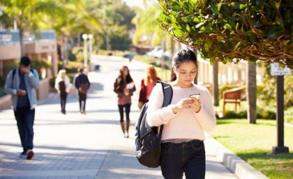 A street in daylight with people with pedestrians looking at their phones as they walk with a young woman in the foreground.